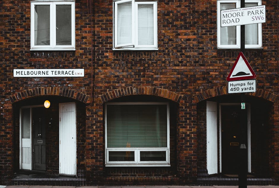 A two-storey brick residential building situated on Melbourne Terrace, featuring white-framed windows with horizontal blinds on the upper level and a large window with closed blinds on the ground level. The building has a dark grey front door positioned beneath a rounded brick archway, with a small, glowing exterior light mounted beside it. To the right, there is another entrance with a dark door also beneath a brick arch, and near this doorway, a mailbox is visible. The building displays two street signs; one in the upper left corner labeled 'Melbourne Terrace' with a small red 'SW6' code, and another in the upper right corner indicating the intersection of Moore Park Road with additional signage for road conditions, including a triangular warning sign about speed humps for 450 yards. Surrounding the building is a paved pavement and a street scene typical of an urban area. The environment is lit with natural daylight, providing a clear view of the brick textures and architectural details, relevant to private properties where waste clearance services like those offered by Waste Clearance Fulham could be employed for on-site rubbish removal or private waste handling.