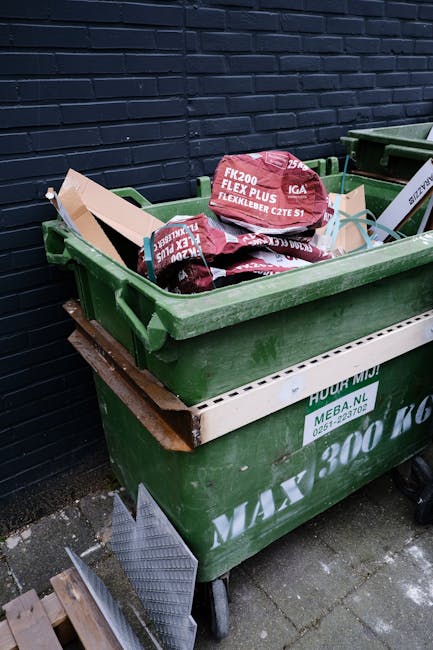 A large green plastic waste bin positioned on a concrete pavement beside a dark brick wall. The bin is filled with black and red bags of garden or landscaping materials, including soil or compost, with visible labels and branding. The open lid displays the bags stacked loosely inside. A smaller white container is attached to the side of the bin, displaying a website URL and contact number for waste collection services. A metal plank and a piece of corrugated metal are resting on the pavement adjacent to the bin, indicating ongoing waste disposal or clearance activities. The environment suggests an outdoor area set up for private rubbish collection, emphasizing the use of self-managed waste removal solutions instead of local authority services. The scene is illuminated by natural light, with some dirt and debris visible on the ground beneath the bin, reflecting typical conditions for a commercial or residential waste disposal setup. Waste Clearance Fulham’s service focus on convenient rubbish removal is subtly supported by the presence of multiple waste containers ready for collection or transport.