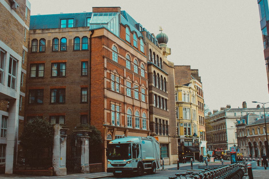 A narrow city street lined with historic multi-story buildings featuring brick facades, arched and rectangular windows, and intricate architectural details. In the foreground, a white refuse collection truck with a modern design and clean exterior is parked along the curb, adjacent to a small section of pavement and street bollards. To the left, a portion of a building with white window frames and a brick wall is visible, while in the background, a mix of older and more ornate architecture, including a building with a rounded turret topped by a green dome and golden cross, dominates the skyline. The street scene is illuminated by natural daylight, with soft shadows cast on the buildings and road, creating a quiet, urban atmosphere. The presence of the waste collection truck and surrounding infrastructure subtly relates to private waste handling and rubbish removal services, fitting the context of alternative rubbish collection options in dense city environments.