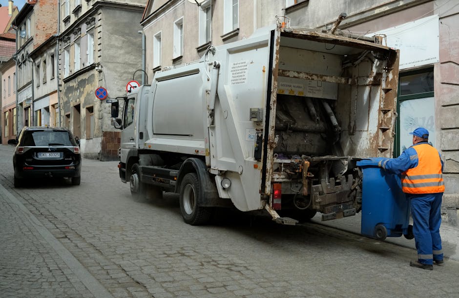 The image shows a white refuse collection vehicle positioned on a narrow cobblestone street in an urban area, with a worker in a bright orange and blue uniform and protective gloves loading a blue wheeled bin into the back of the truck. The truck's rear compartment, which appears partially rusted and has an open hatch, is designed for compacting waste materials. In the foreground, a black car is parked on the street adjacent to the truck, while in the background, residential buildings with multicoloured facades and tall windows line the street. Two circular signs are mounted on a pole attached to one of the buildings, indicating parking and no-parking zones. The street scene captures a routine waste collection process that aligns with private on-site clearance and independent rubbish removal services, often preferred for speed and convenience in urban environments. The scene is well-lit with natural daylight, providing clear visibility of textures such as the rough cobblestones, the smooth surface of the truck, and the worker's safety gear, emphasizing professional waste management activity.