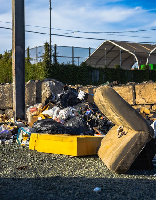 An overflowing collection of mixed waste and rubbish bags, including black, white, and clear plastic bags, cardboard boxes, and discarded packaging materials, is piled on a paved urban sidewalk next to a metal railing. The waste appears to consist of paper, cardboard, plastic containers, and miscellaneous household items. Behind the pile, there is a grey waste collection bin with a blue lid marked “mixed paper & card,” which is visibly full with some waste spilling over the top. To the left, a small grey car is parked adjacent to the waste area, with a UK license plate visible. In the background, there are storefronts with yellow and black signage, and a building with scaffolding and building wrap indicating ongoing construction or refurbishment. The scene is lit with natural daylight, and the general environment suggests a typical city street where private rubbish disposal or skip alternatives are often used, which Waste Clearance Fulham provides as part of their rubbish removal services. The image highlights the necessity for efficient waste collection solutions within urban settings.