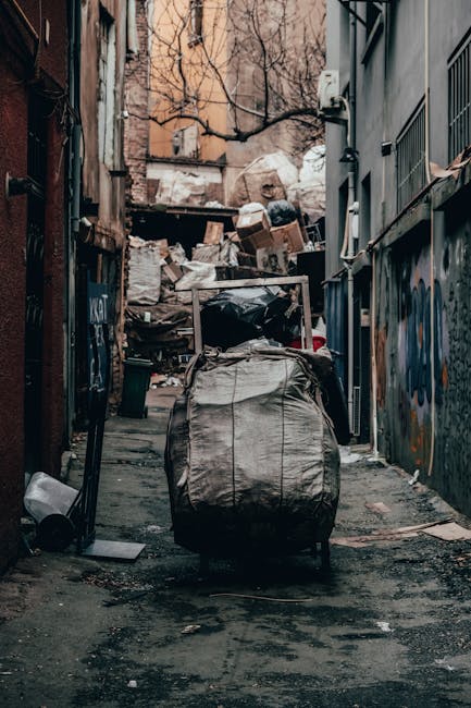 A narrow urban alleyway cluttered with overflowing black rubbish bags and cardboard boxes piled against a wall in the background. The bags are made of thick, crinkled plastic, showing signs of wear with some tearing. The ground is uneven, stained, and scattered with smaller debris, suggesting recent rubbish collection or ongoing clearance. On the right side, there is a metal railing attached to the side of a building, with graffiti visible on the wall beneath. On the left, a weathered brick wall features a few discarded items, including a plastic container near the ground. The alley is partially shaded, with faint light filtering through leafless tree branches overhead and the surrounding buildings, creating a dull, utilitarian atmosphere. This scene illustrates a typical example of private waste disposal and clearance often handled by waste removal services such as Waste Clearance Fulham, supporting alternative waste management outside local authority collection methods.