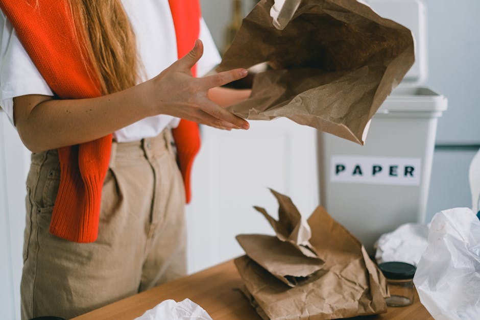 The image shows a person wearing a white shirt and beige trousers, with part of an orange-red knitted cardigan visible, throwing crumpled brown paper into a white recycling bin labeled 'PAPER.' The person's hands are mid-motion, holding a large crumpled paper bag, with additional crumpled paper and white plastic waste on the wooden table in front. In the background, the recycling container is placed against a wall, emphasizing the context of waste sorting and private rubbish disposal, relevant to independent rubbish clearance services for residential properties. The environment appears well-lit with natural or soft indoor lighting, indicating an indoor setting dedicated to waste management or recycling activities, supporting the overall theme of rubbish removal and responsible waste handling on behalf of waste clearance providers.