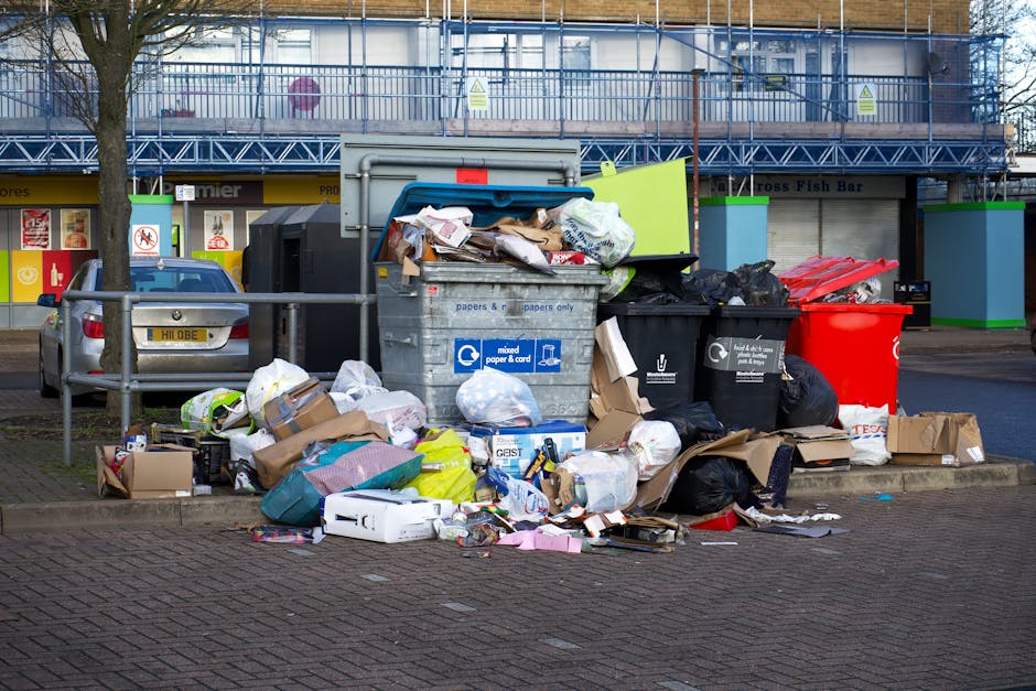 A narrow urban alleyway cluttered with overflowing black rubbish bags and cardboard boxes piled against a wall in the background. The bags are made of thick, crinkled plastic, showing signs of wear with some tearing. The ground is uneven, stained, and scattered with smaller debris, suggesting recent rubbish collection or ongoing clearance. On the right side, there is a metal railing attached to the side of a building, with graffiti visible on the wall beneath. On the left, a weathered brick wall features a few discarded items, including a plastic container near the ground. The alley is partially shaded, with faint light filtering through leafless tree branches overhead and the surrounding buildings, creating a dull, utilitarian atmosphere. This scene illustrates a typical example of private waste disposal and clearance often handled by waste removal services such as Waste Clearance Fulham, supporting alternative waste management outside local authority collection methods.